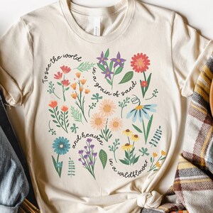 May include: A beige t-shirt with a floral design and the text "To see the world in a grain of sand and heaven in a wildflower".
