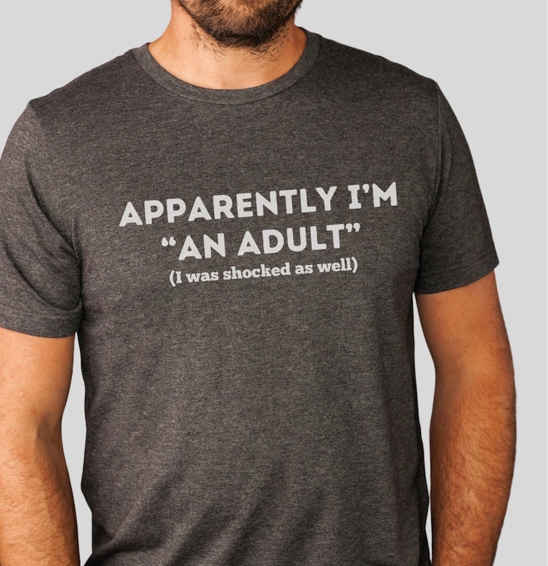 May include: A dark grey t-shirt with the text "APPARENTLY I'M "AN ADULT" (I was shocked as well)" printed in white.