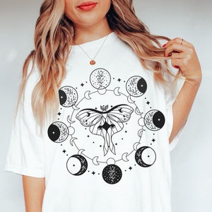 May include: White t-shirt with a black and white graphic design of a moth with wings spread, surrounded by a circle of crescent moons and stars.