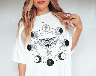 May include: White t-shirt with a black and white graphic design of a moth with wings spread, surrounded by a circle of crescent moons and stars.