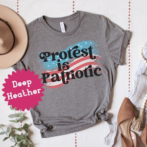 Puede incluir: Una camiseta gris con el texto "Protest is Patriotic" en una fuente retro. El texto est&aacute; superpuesto a un gr&aacute;fico de bandera estadounidense roja, blanca y azul. La camiseta se muestra sobre una superficie blanca con un sombrero, pantalones vaqueros y zapatos.