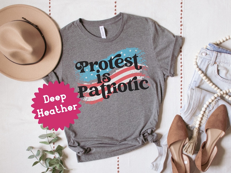 Puede incluir: Una camiseta gris con el texto "Protest is Patriotic" en una fuente retro. El texto est&aacute; superpuesto a un gr&aacute;fico de bandera estadounidense roja, blanca y azul. La camiseta se muestra sobre una superficie blanca con un sombrero, pantalones vaqueros y zapatos.