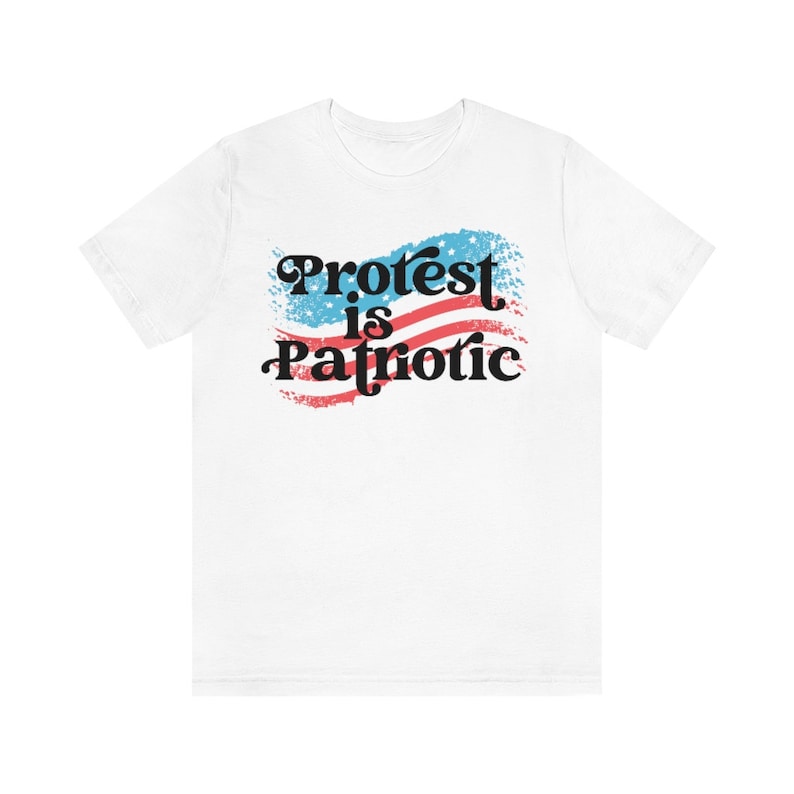Puede incluir: Camiseta blanca con un dise&ntilde;o de bandera estadounidense roja, blanca y azul y el texto "Protest is Patriotic" en negro.