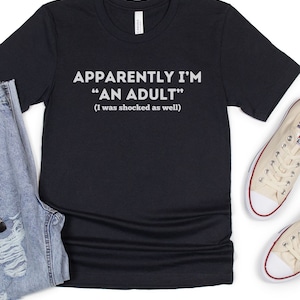 May include: Black t-shirt with white text that reads "Apparently I'm "An Adult" (I was shocked as well)"