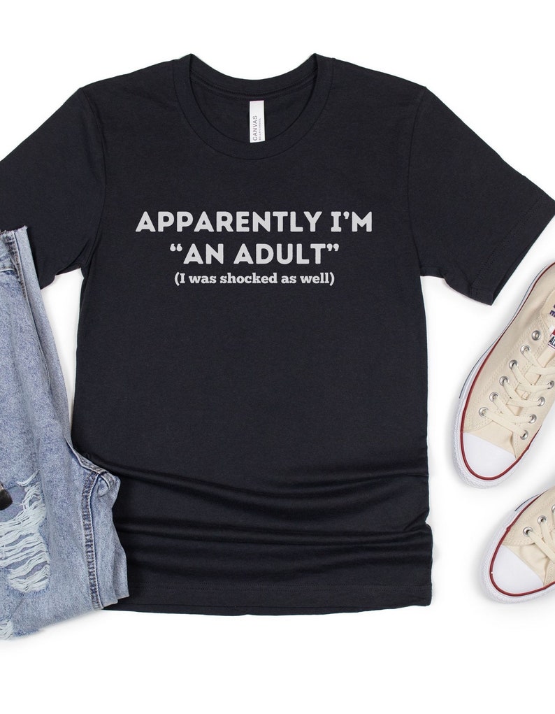 May include: Black t-shirt with white text that reads "Apparently I'm "An Adult" (I was shocked as well)"