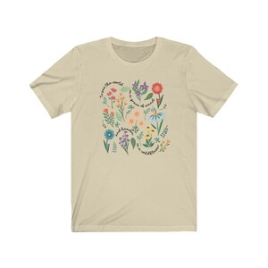 May include: A beige t-shirt with a floral design and the text "To see the world, we are a grain of sand, and heaven, we are a wildflower."