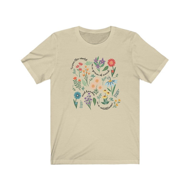 May include: A beige t-shirt with a floral design and the text "To see the world, we are a grain of sand, and heaven, we are a wildflower."