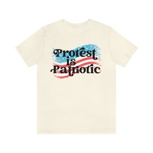 Puede incluir: Una camiseta beige con un gr&aacute;fico de una bandera estadounidense con el texto "Protest is Patriotic" en negro.