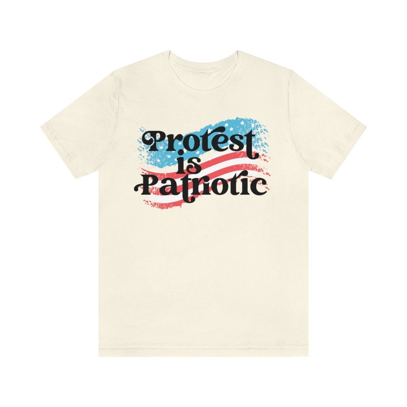 Puede incluir: Una camiseta beige con un gr&aacute;fico de una bandera estadounidense con el texto "Protest is Patriotic" en negro.