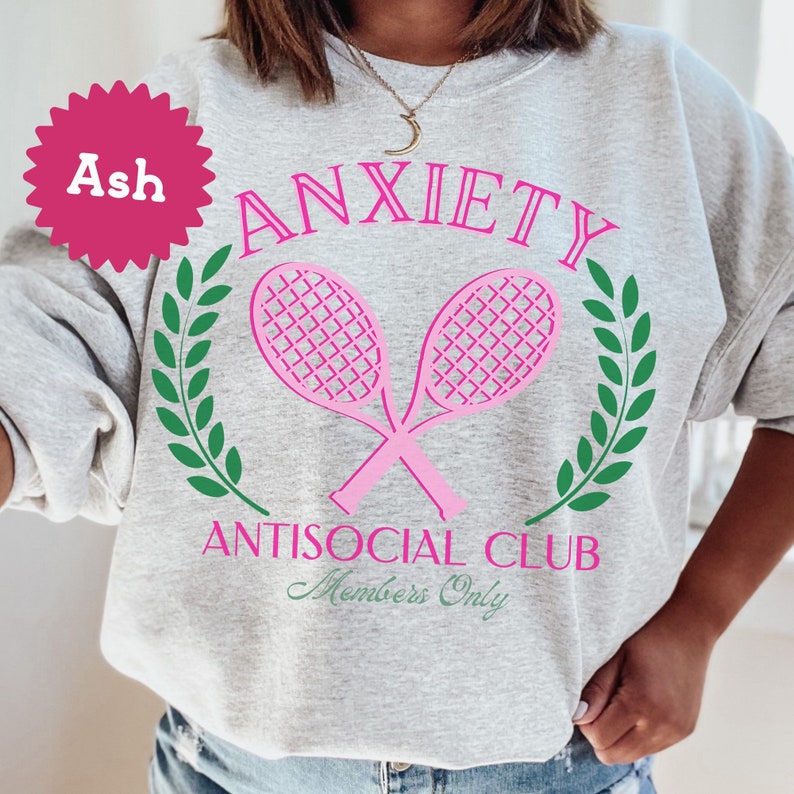May include: Ash grey sweatshirt with a pink graphic design. The graphic features two pink tennis rackets crossed over each other with green laurel wreaths on either side. The text "Anxiety Antisocial Club Members Only" is printed below the rackets.