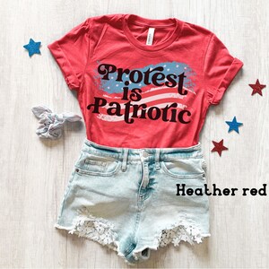 Puede incluir: Camiseta roja con un dise&ntilde;o de bandera estadounidense blanca y azul y el texto "Protest is Patriotic". La camiseta est&aacute; sobre una superficie de madera blanca con estrellas azules y rojas.
