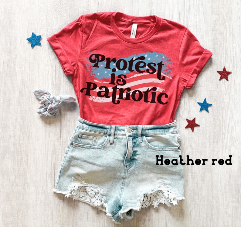 Puede incluir: Camiseta roja con un dise&ntilde;o de bandera estadounidense blanca y azul y el texto "Protest is Patriotic". La camiseta est&aacute; sobre una superficie de madera blanca con estrellas azules y rojas.