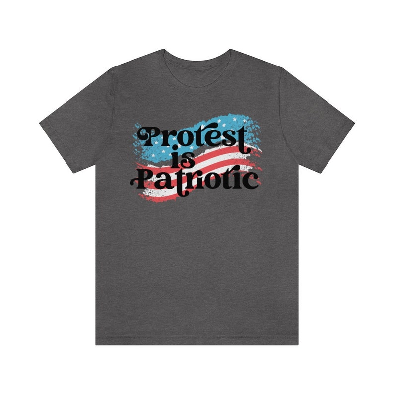 Puede incluir: Una camiseta gris oscuro jaspeada con un dise&ntilde;o de bandera estadounidense desgastado y el texto "Protest is Patriotic" en negro.