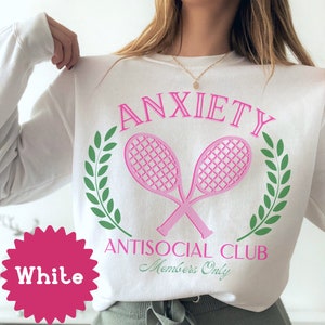 May include: White sweatshirt with pink text that reads "Anxiety Antisocial Club Members Only" with a pink tennis racket graphic and green laurel wreaths.
