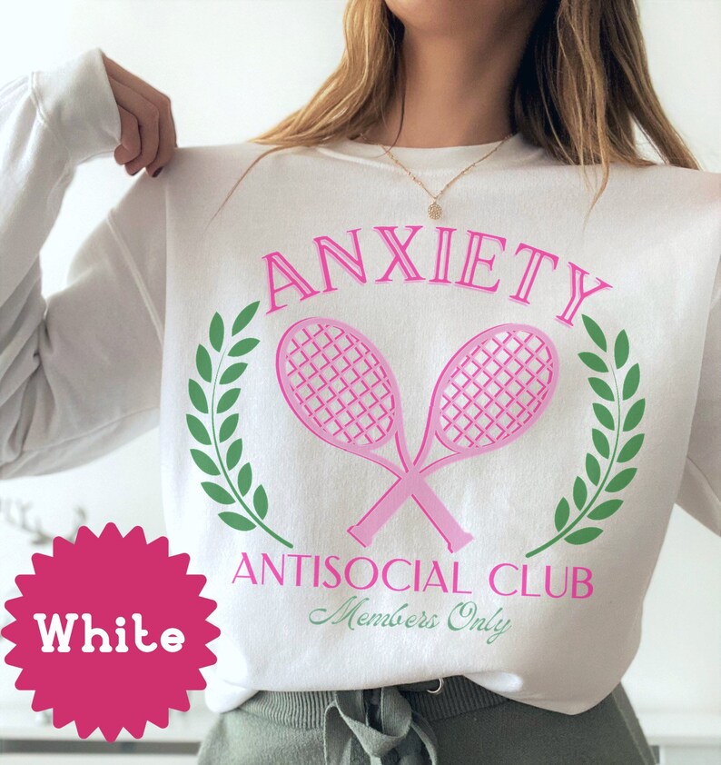 May include: White sweatshirt with pink text that reads "Anxiety Antisocial Club Members Only" with a pink tennis racket graphic and green laurel wreaths.