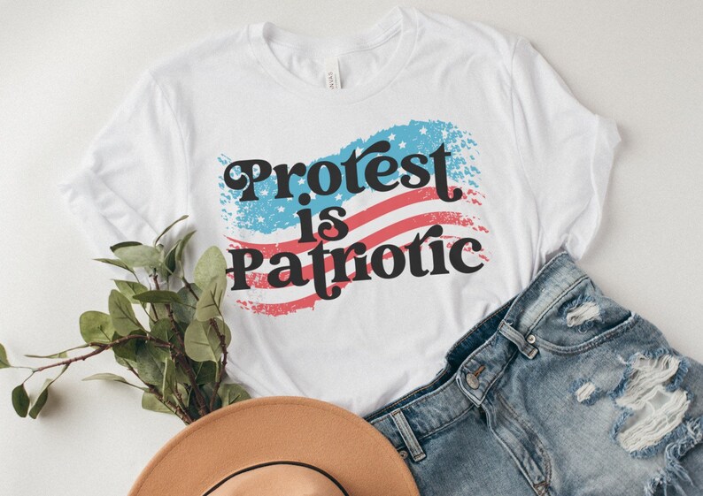 Puede incluir: Camiseta blanca con un dise&ntilde;o de bandera estadounidense desgastado y el texto "Protest is Patriotic" en negro.