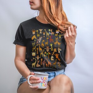 Pu&ograve; includere: Una maglietta nera con un motivo di molte giraffe colorate. La persona che indossa la maglietta tiene una tazza bianca con il testo "You got this" in lettere colorate.