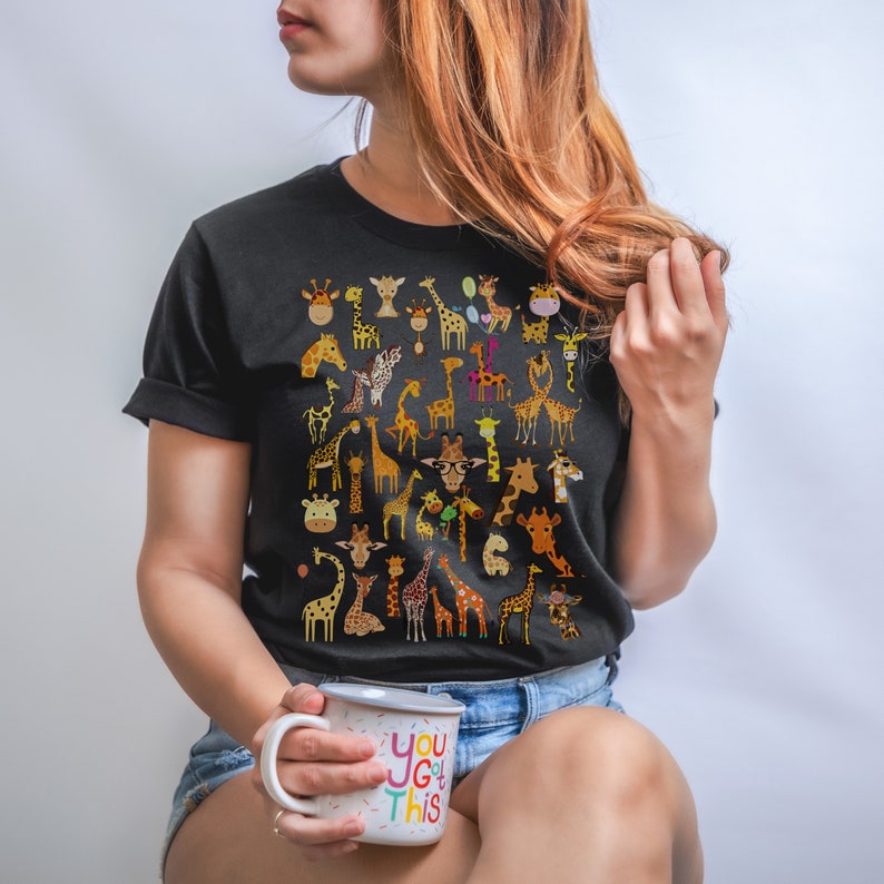Pu&ograve; includere: Una maglietta nera con un motivo di molte giraffe colorate. La persona che indossa la maglietta tiene una tazza bianca con il testo "You got this" in lettere colorate.
