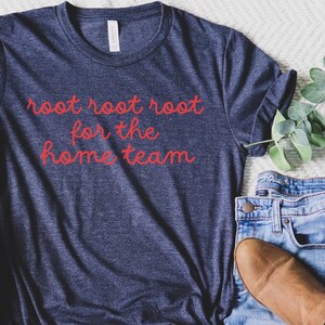 May include: A navy blue t-shirt with the red text "root root root for the home team." The shirt is folded, and placed next to blue jeans and a brown boot. The shirt is made of a soft material.