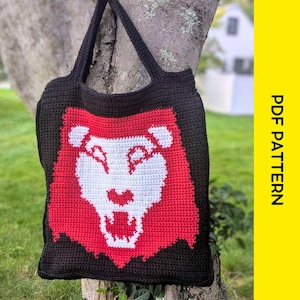 Puede incluir: Bolso tote de crochet negro con un diseño de cara de león rojo y blanco. El bolso tiene dos asas negras y está colgado de un árbol. Las palabras "PDF PATTERN" están impresas verticalmente sobre un fondo amarillo.