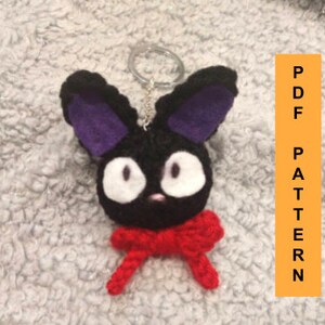 May include: Black felt keychain with purple ears, white eyes, and a red crocheted bow. The keychain is shaped like a cat.