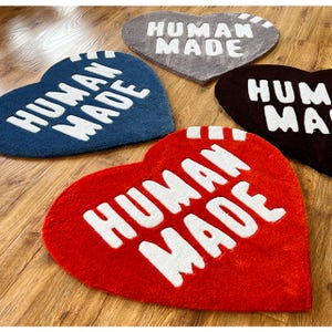 May include: Four heart-shaped rugs with the text "HUMAN MADE" in white lettering. The rugs are in red, blue, brown, and gray.