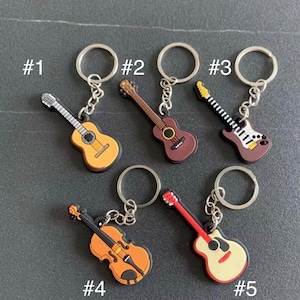 May include: A collection of five musical instrument keychains. The keychains feature miniature guitars, a violin, and an electric guitar in various colors, including brown, yellow, red, and white. Each keychain has a silver ring for attaching keys.