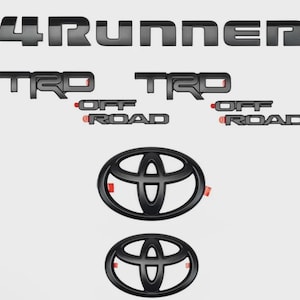 May include: A set of matte black automotive emblems including one 4RUNNER badge, two TRD OFF ROAD badges, and two Toyota oval logos. Each piece features a clean, bold typeface and design, with small red adhesive tabs visible on the sides.