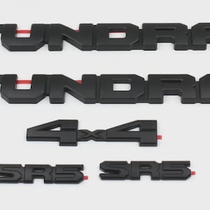 May include: A set of matte black automotive emblems including two TUNDRA nameplates, one 4x4 emblem, and two SR5 badges. Each piece features a bold, block-letter design with visible red adhesive backing strips on the reverse side of the letters.