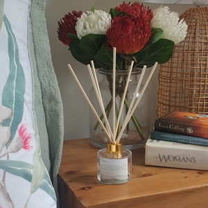 May include: A glass diffuser bottle with wooden reeds and a gold-coloured cap is on a wooden bedside table. The bottle has a label with text. A bouquet of red and white flowers is in a glass vase. Books and a woven lamp are also on the table.