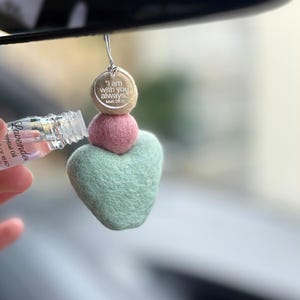 May include: A hanging car air freshener composed of three felted wool spheres in beige, pink, and light green, with the beige sphere featuring the printed text I am with you always. Matt 28:20. A small glass bottle of lavender essential oil is nearby.