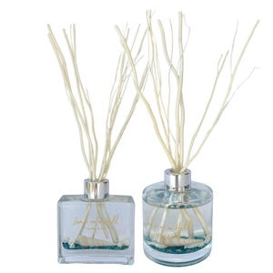 May include: Two glass reed diffusers with silver tops. One is square, the other round. Both contain seashells and light blue liquid. The diffusers have light-coloured reeds and the text "Lamps on a Hill" on the front.