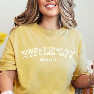 May include: A woman wearing a yellow t-shirt with the text "Hufflepuff Hogwarts" in white. She is sitting on a white couch with a yellow pillow and holding a pink mug with white flowers.
