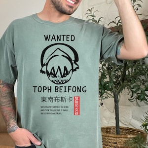 May include: A green t-shirt with a black graphic of a cartoon character with the text "WANTED TOPH BEIFONG 東南布斯卡 SHE DISGUISES HERSELF AS BLIND AND EVEN THOUGH SHE IS SMALL SHE IS VERY DANGEROUS 危險的女孩"