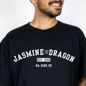 Pode incluir: Camiseta preta com texto branco que diz "JASMINE DRAGON" em arco na parte superior, com "EST. 2006" e "BA SING SE" abaixo. A camiseta é azul marinho escuro e tem mangas curtas.