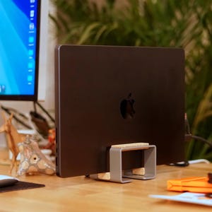 Puede incluir: Un ordenador portátil negro con el logotipo de Apple está apoyado en un soporte de metal gris con detalles de corcho. El soporte está sobre un escritorio de madera. Un monitor de ordenador y vegetación son visibles en el fondo.
