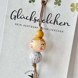 Könnte beinhalten: Ein Holzpuppenanhänger mit einem lächelnden Gesicht, blonden Haaren und einem roten Rouge. Unter dem Gesicht befindet sich eine blau-weiß gestreifte Perle. Der Anhänger hängt an einer braunen Schnur. Der Hintergrund enthält Text auf Deutsch.