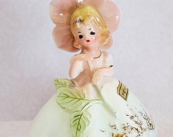 May include: A porcelain figurine of a girl wearing a pink flower hat and a light blue dress with green leaves and gold accents. The figurine is labeled "Night."