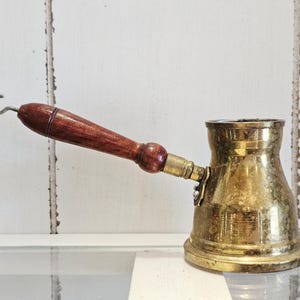 May include: A brass coffee pot with a wide base and a narrow neck, featuring a long, dark brown wooden handle with a small metal hook at the end. The brass surface shows a weathered, metallic patina with visible signs of age and use.