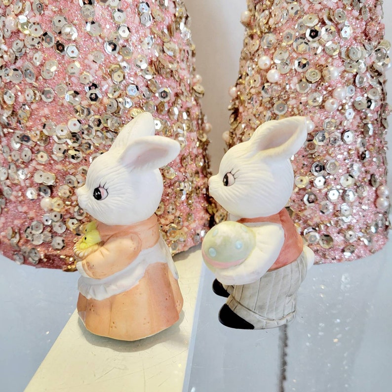 Puede incluir: Dos figuritas de conejo de porcelana blanca con detalles pintados. Un conejo sostiene una cesta de pollitos amarillos y el otro sostiene un huevo de Pascua pastel. Los conejos est&aacute;n de pie sobre una superficie blanca.