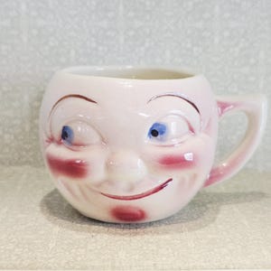 May include: A novelty ceramic mug featuring a smiling face. The mug is pale pink with blue eyes, red cheeks, and a red mouth. It has a handle and is designed for holding and drinking beverages.
