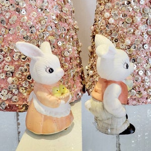 Puede incluir: Dos figuritas de conejo de cer&aacute;mica blanca. Un conejo sostiene una cesta de pollitos amarillos y lleva un vestido rosa. El otro conejo lleva un chaleco rojo y pantalones grises.