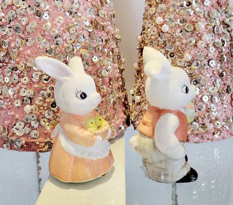 Puede incluir: Dos figuritas de conejo de cer&aacute;mica blanca. Un conejo sostiene una cesta de pollitos amarillos y lleva un vestido rosa. El otro conejo lleva un chaleco rojo y pantalones grises.