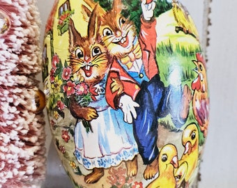 May include: A vintage Easter egg made of paper mache with a colorful illustration of two bunnies and a rooster. The bunnies are dressed in red and blue clothing and are holding flowers. The egg is decorated with a green, yellow, and blue background.