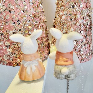 Puede incluir: Dos figuritas de conejo de cer&aacute;mica blanca con detalles pintados. Un conejo lleva un vestido rosa con un delantal blanco y el otro lleva un chaleco color melocot&oacute;n y pantalones grises.
