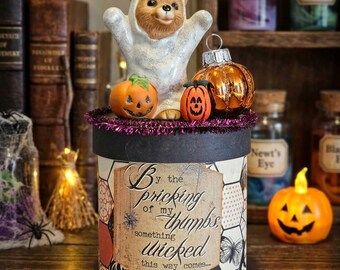 May include: A cylindrical box with a black lid and a label reading By the pricking of my thumbs something wicked this way comes. The lid features a small bear figurine in a white ghost costume, two miniature pumpkins, and a metallic orange ornament.