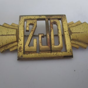 May include: A gold-toned metal brooch with a square frame and the letters "ZGD" inside. The frame is decorated with two art deco-style wings on either side.