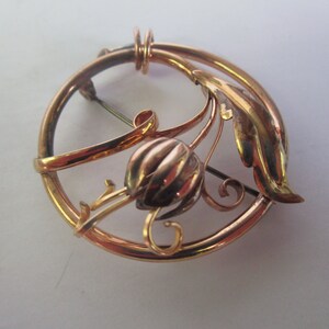 May include: A gold-toned brooch with a stylized floral design. The brooch features a circular frame with a delicate leaf and flower motif.