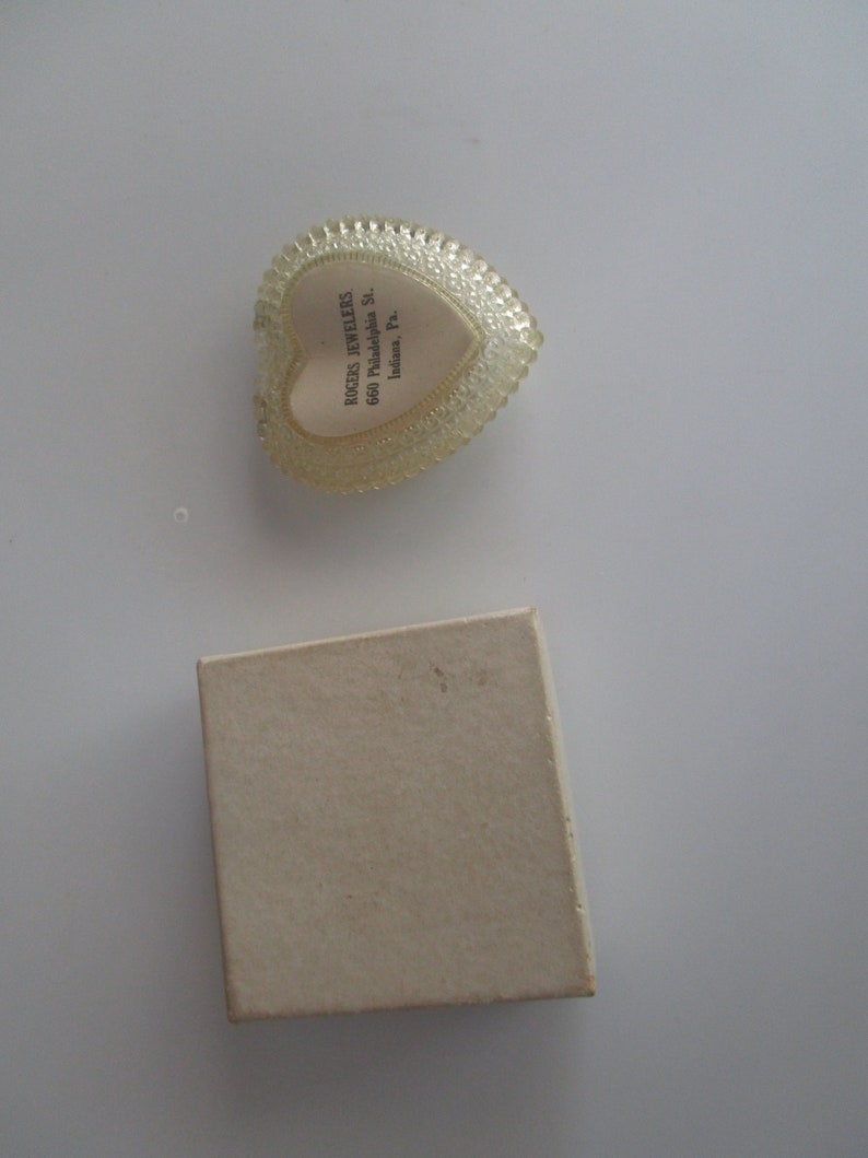 May include: A heart-shaped, clear plastic jewellery box with a scalloped edge and a white interior. The box has the text "ROGERS JEWELERS 660 Philadelphia St. Indiana, Pa." A small, beige cardboard box is also present.