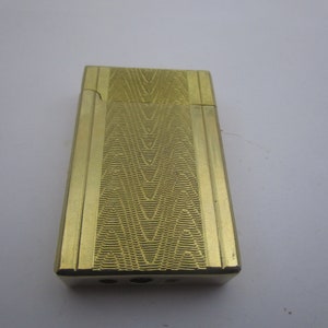 May include: A gold-colored lighter with a textured, geometric pattern. The lighter has a rectangular shape and a slightly rounded top.
